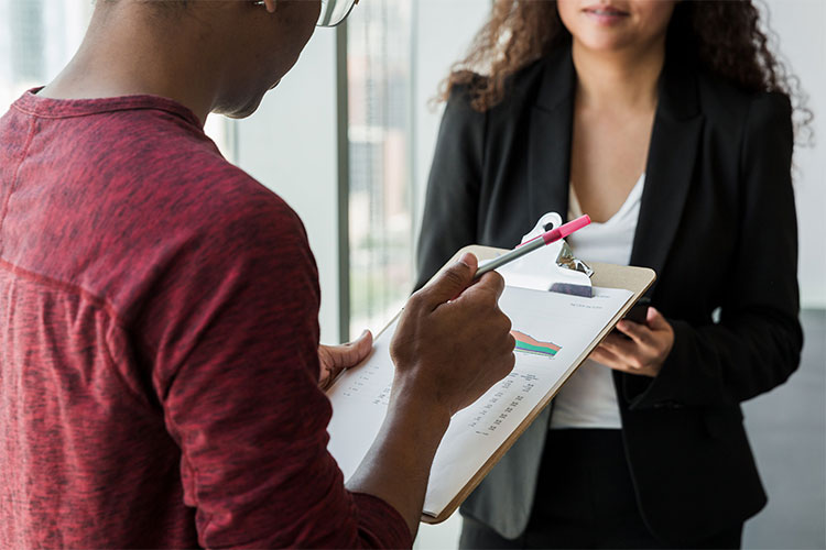 A person holds a clipboard a pen, and is standing in front of a second person, facing eachother.